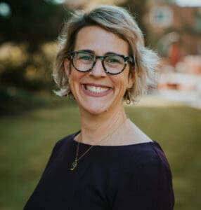 A woman with short blonde hair, glasses, and a dark top is smiling outdoors with blurred greenery and buildings in the background.