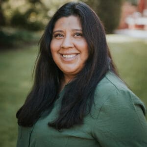 A woman with long dark hair wearing a green shirt smiles outdoors, with greenery and sunlight in the background.
