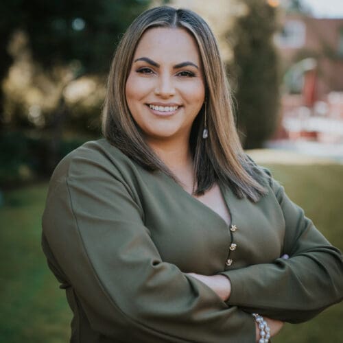 Woman with straight, shoulder-length hair and olive green blouse stands outdoors with arms crossed, smiling at the camera.