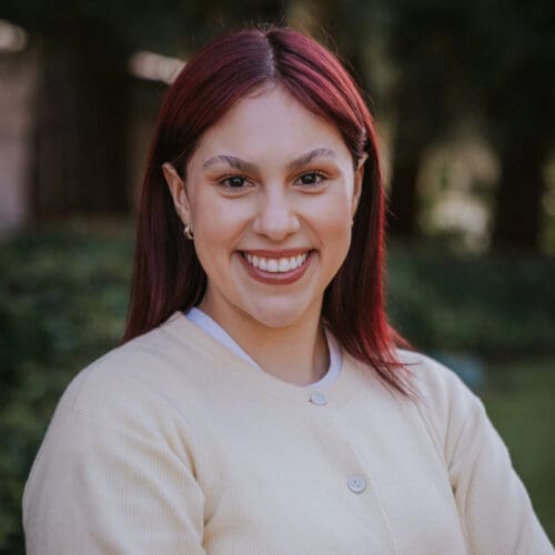 A woman with straight, shoulder-length red hair smiles at the camera, standing outdoors in front of greenery and trees, wearing a light beige buttoned shirt.