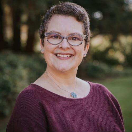 A person with short hair and glasses, wearing a maroon sweater and a pendant necklace, smiles outdoors with greenery in the background.